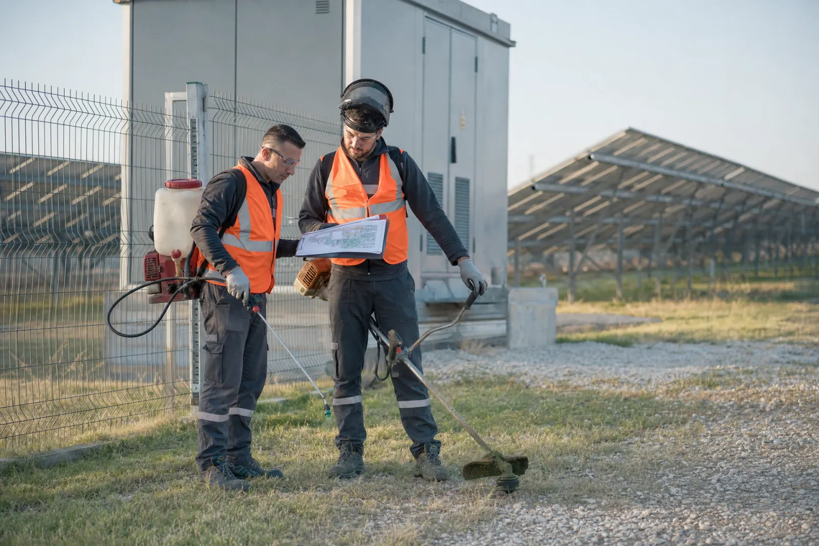 Turuncu yelekli iki saha çalışanı, GES trafo binası önünde sırt pompası ve tırpanla çalışma planına bakıyor
